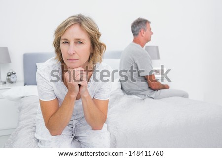 Thoughtful couple sitting on different sides of bed having a dispute with woman looking at camera in bedroom at home