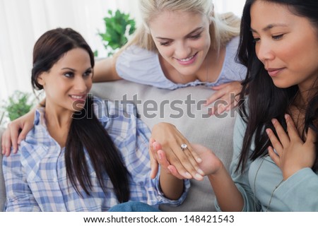Smiling friends admiring brunettes engagement ring at home on couch