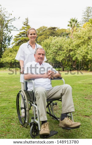 Happy man sitting in a wheelchair with his nurse pushing him smiling at camera in the park