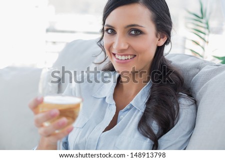 Cheerful pretty brunette drinking white wine sitting on sofa in her living room