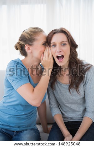 Astonished woman being told a secret by her friend and sitting on the sofa