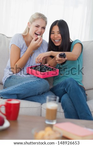 Friends laughing at tv and sharing box of chocolates at home on couch