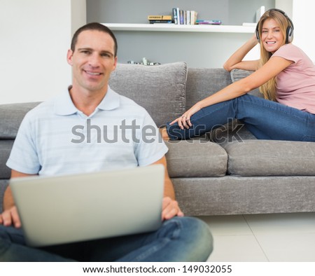 Man sitting on floor using laptop with woman listening to music on the sofa both looking at camera in sitting room at home
