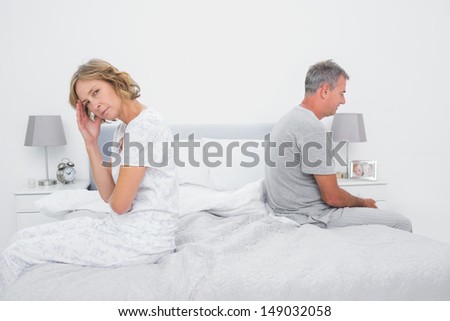 Annoyed Couple Sitting On Different Sides Of Bed Having A Dispute With Woman Looking At Camera In Bedroom At Home