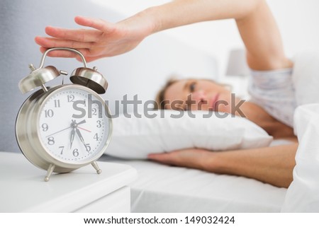 Tired blonde woman turning off her alarm in the morning at home in bedroom