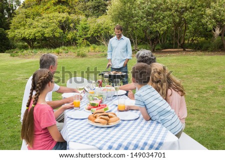 Cheerful extended family sitting at picnic table watching father at the barbecue