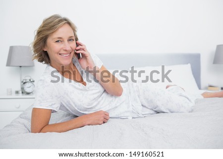 Content blonde woman lying on bed making a phone call smiling at camera in bedroom at home