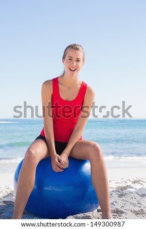 Fit happy woman sitting on exercise ball looking at camera on the beach