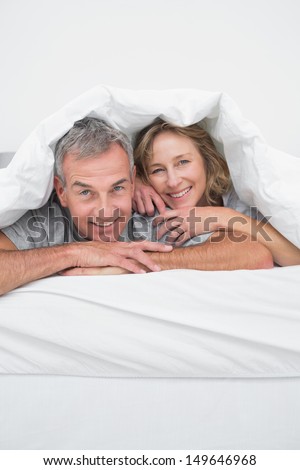 Cheerful middle aged couple under the duvet smiling at camera in bedroom at home
