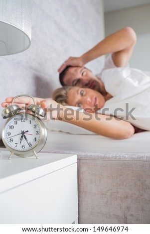 Annoyed couple looking at alarm clock in the morning with woman turning it off at home in bedroom