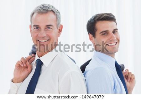Smiling businessmen in bright office posing back to back together while holding their jacket