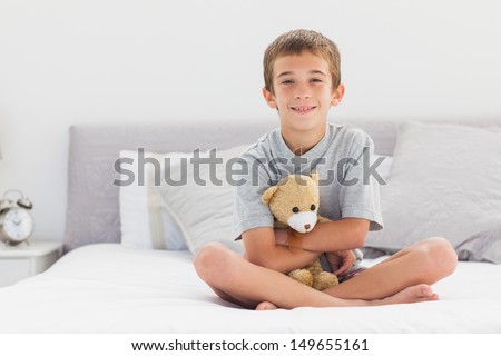 Smiling little boy sitting on bed holding his teddy bear at home