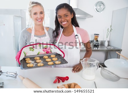 Cheerful woman showing freshly baked cookies with friend looking at camera in kitchen