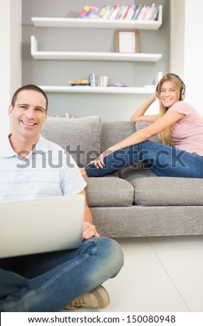 Man sitting on floor using laptop with woman listening to music on the sofa both smiling at camera in sitting room at home