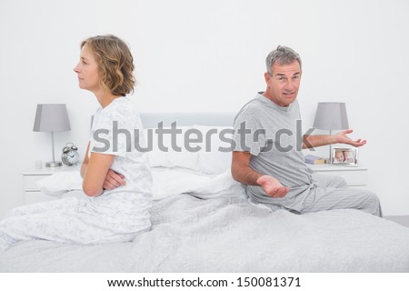 Couple sitting on different sides of bed having a dispute with husband gesturing at camera in bedroom at home