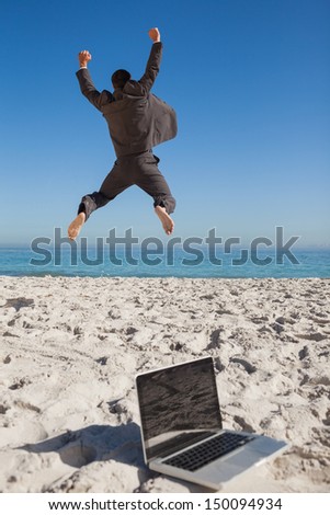 Victorious businessman in suit jumping leaving his laptop on the beach