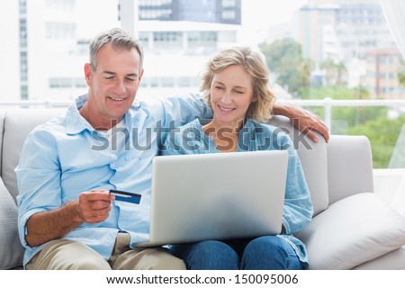 Smiling Couple Sitting On Their Couch Using The Laptop To Buy Online At Home In The Sitting Room