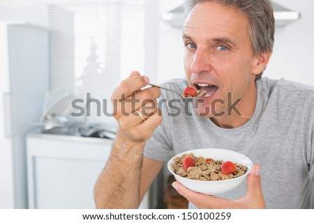 Happy Man Eating Cereal For Breakfast In Kitchen Looking At Camera