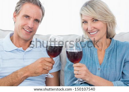 Couple toasting with red wine on the sofa smiling at camera at home in living room