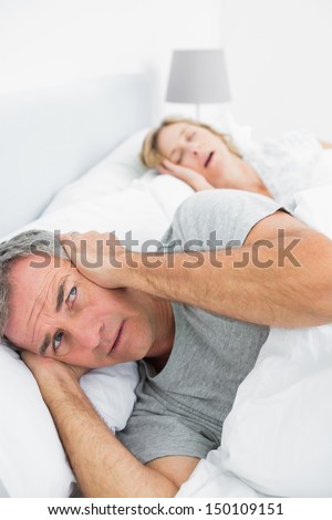 Irritated man blocking his ears from noise of wife snoring at home in bedroom