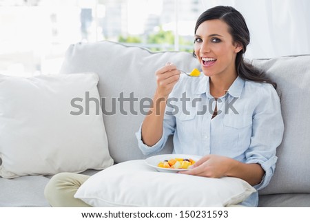 Happy woman sitting on the sofa eating fruit salad in her living room