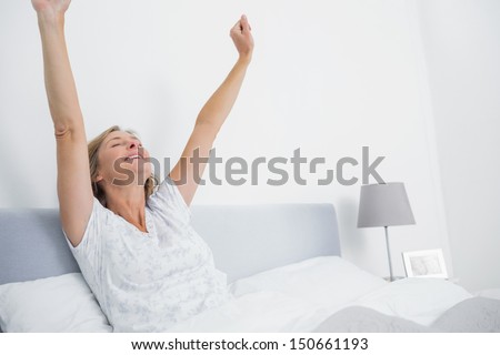 Well rested blonde woman stretching in bed and smiling in bedroom at home