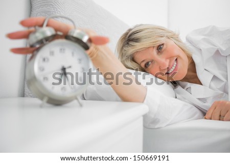 Blonde Woman Turning Off Ringing Alarm Clock Lying In Bed At Home