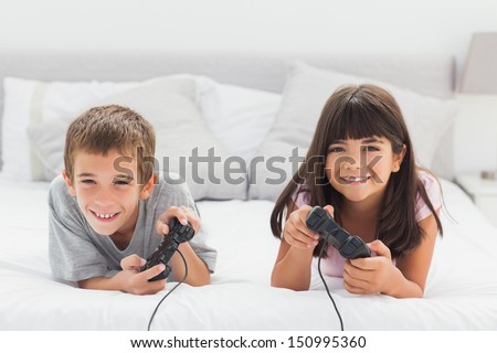 Smiling Siblings Lying On Bed Playing Video Games Together At Home