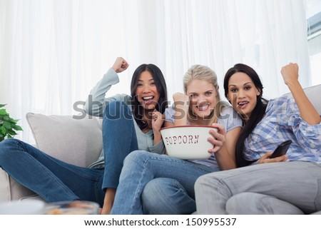 Friends cheering at television with bowl of popcorn at home on couch