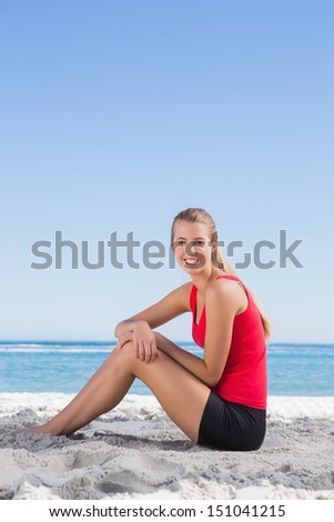 Athletic blonde sitting on sand smiling at camera on the beach
