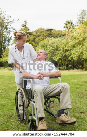 Happy man sitting in a wheelchair talking with his nurse pushing him at the park on sunny day