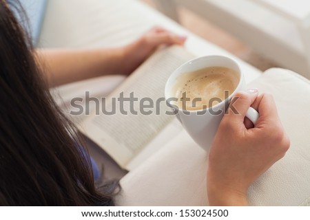 Woman lying on the sofa reading a book holding her coffee in living room at home