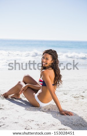 Sexy tanned woman in bikini smiling at camera while sitting on the beach