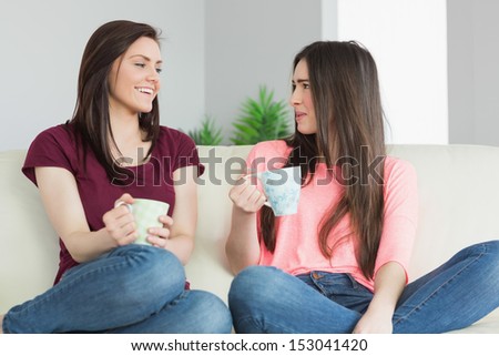 Two Teenage Girls Sitting On A Sofa In A Living Room Having A Conversation And Holding A Beverage