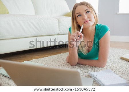 Thoughtful blonde lying on floor doing her assigment using laptop in living room at home