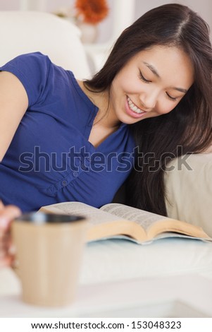 Relaxed asian girl lying on the sofa reading a novel and drinking hot beverage in living room at home