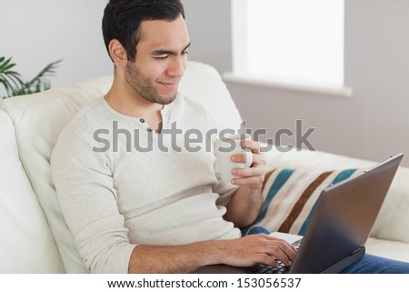 Calm Attractive Man Drinking Coffee While Working On His Laptop In Bright Living Room