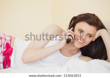Smiling brunette calling someone with a mobile phone lying on a bed in a bedroom