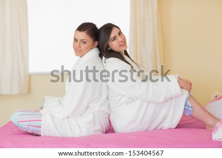 Two friends wearing bathrobes sitting back to back smiling at camera at home in bedroom
