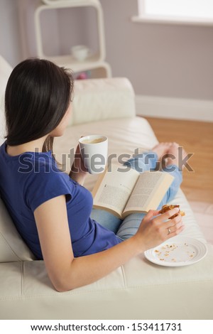 Young woman reading a book and eating pastry with coffee in sitting room at home