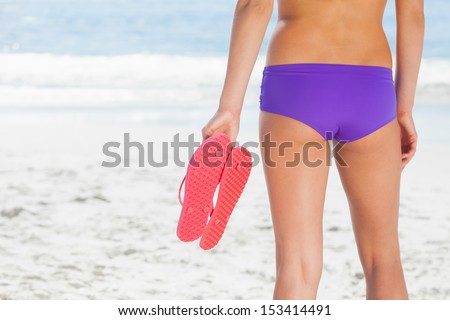 Woman in bikini on beach holding flip flops standing back to camera