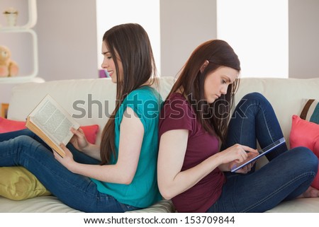 Two happy friends sitting back to back on the sofa reading book and using tablet at home in living room