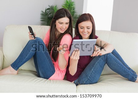 Two happy teenage girls sitting back to back on a sofa using a tablet pc and a mobile phone