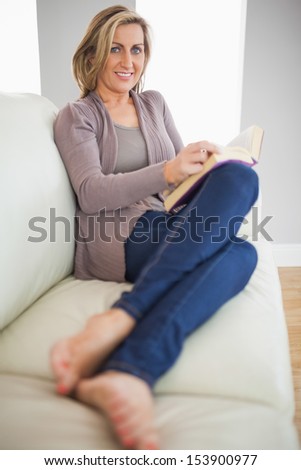 Smiling blonde woman turning pages of a book lying on a sofa in a living room