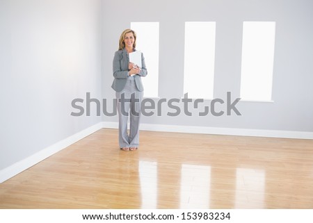 Joyful blonde realtor standing in an empty room holding documents