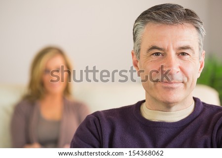 Smiling Mature Man Looking At Camera With His Wife Blurred In The Background