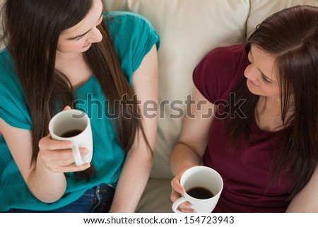 Two smiling friends having coffee and chatting on the couch at home in the living room