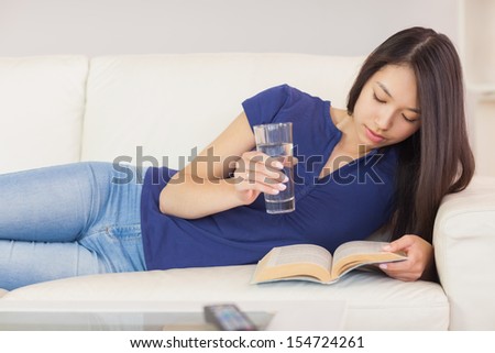 Happy asian girl lying on the sofa reading a novel and drinking water at home in the sitting room
