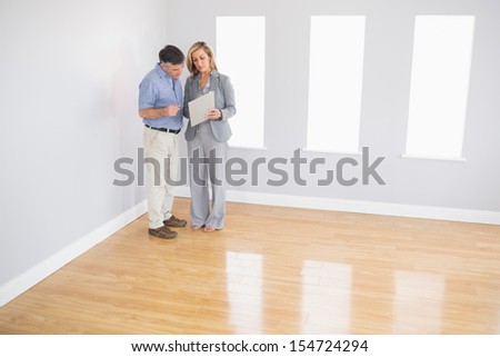 Serious blonde realtor showing an empty room and some documents to a potential mature buyer