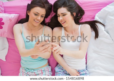 Cheerful friends in pajamas looking at smartphone on bed in bedroom at home